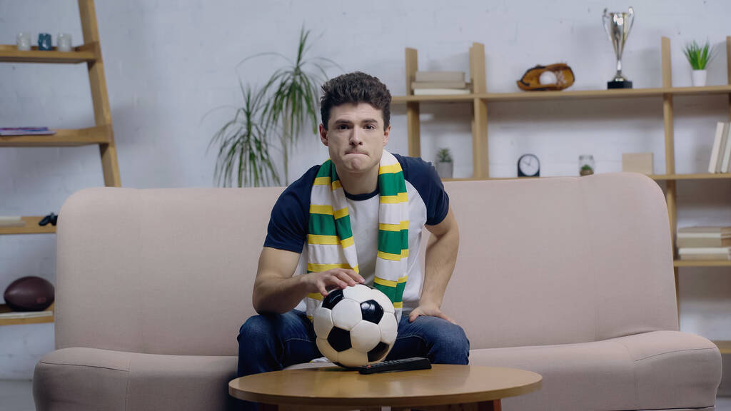 nervous sport fan in striped scarf watching football match near ball on coffee table - Photo, Image