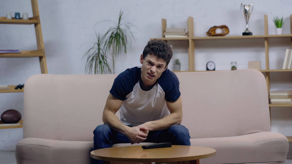 young dissatisfied man watching tv while sitting on sofa near coffee table - Photo, Image
