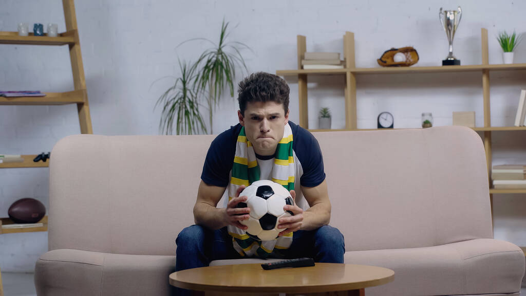 nerveuze sport fan in gestreepte sjaal holding bal tijdens het kijken naar voetbal wedstrijd op home tv - Foto, afbeelding