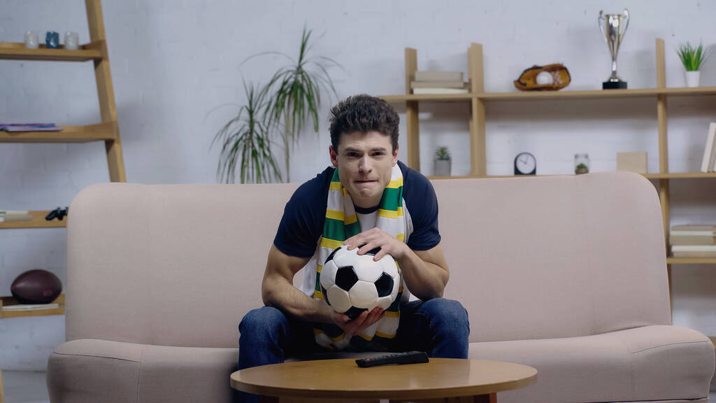 tenso fanático del fútbol sosteniendo pelota de fútbol mientras ve el campeonato en el sofá en casa - Foto, imagen