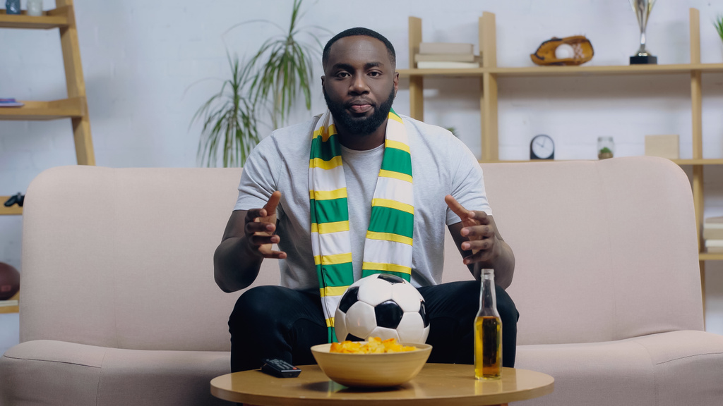 bezorgd Afrikaans Amerikaans voetbal fan gebaren tijdens het kijken naar wedstrijd op tv in de buurt van voetbal, bier en chips - Foto, afbeelding