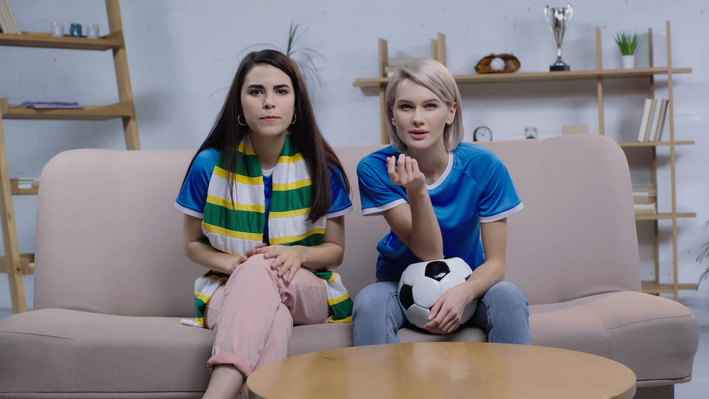 Endişeli esmer ve sarışın kadınlar evdeki kanepede futbol maçı izliyorlar. - Fotoğraf, Görsel
