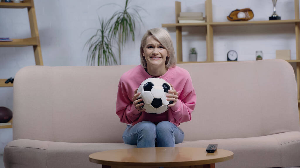 беспокойная фанатка спорта женщина держит футбольный мяч во время просмотра игры по телевизору дома - Фото, изображение