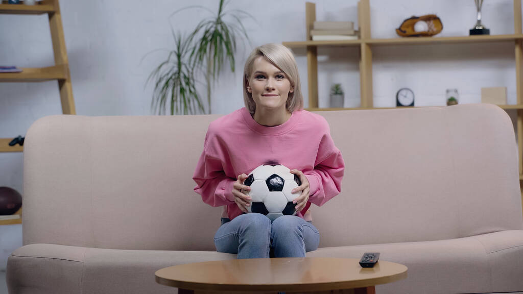 usmívající se fotbalový fanoušek žena drží míč při sledování mistrovství na pohovce doma - Fotografie, Obrázek