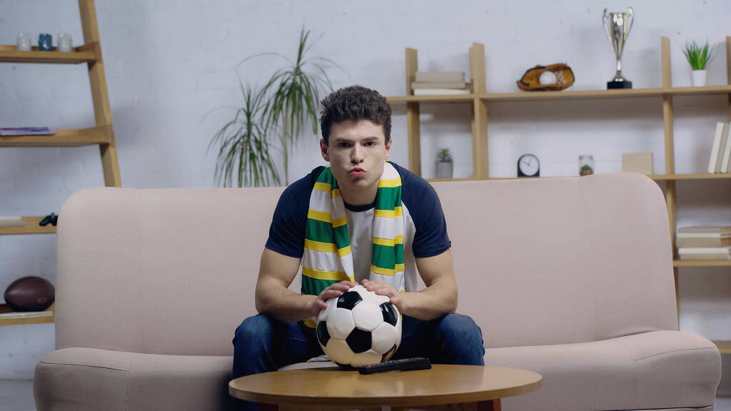 ενθουσιασμένος νεαρός οπαδός του ποδοσφαίρου κάθεται στον καναπέ κοντά μπάλα ποδοσφαίρου στο τραπέζι του καφέ και βλέποντας το παιχνίδι στην τηλεόραση - Φωτογραφία, εικόνα