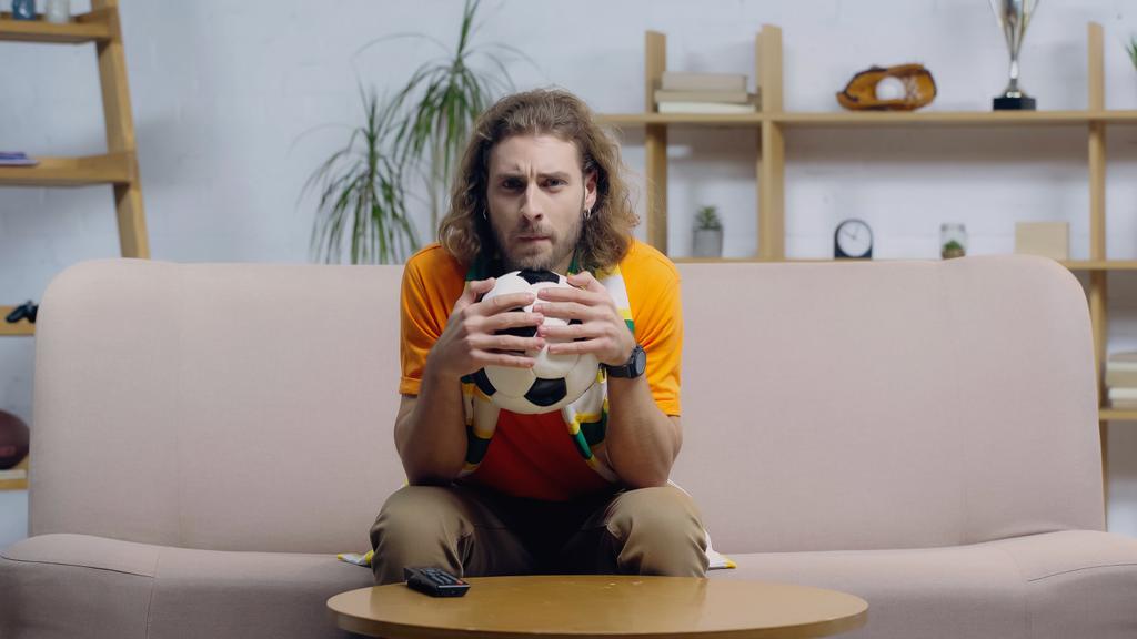 tense man holding soccer ball while watching football match on tv at home - Photo, Image