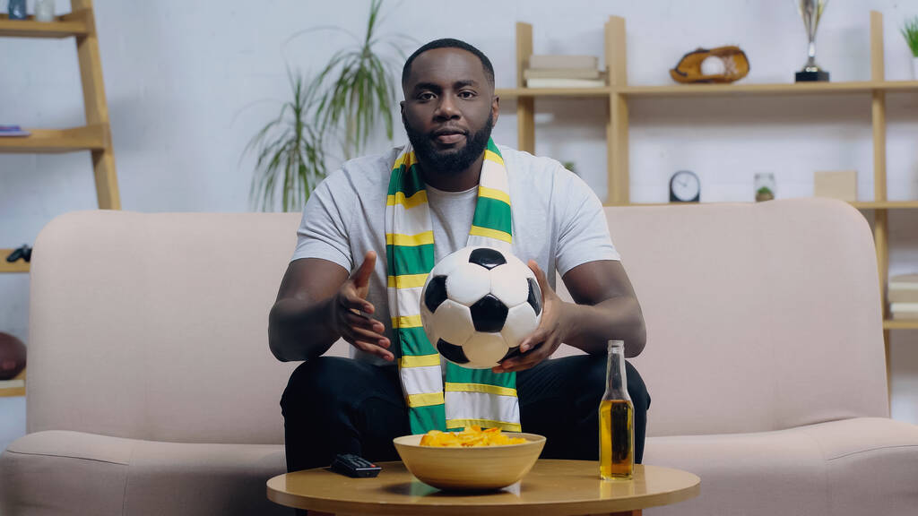 Αφροαμερικάνος με ριγέ φουλάρι που κρατάει μπάλα ποδοσφαίρου ενώ βλέπει πρωτάθλημα κοντά σε μάρκες και μπύρες. - Φωτογραφία, εικόνα