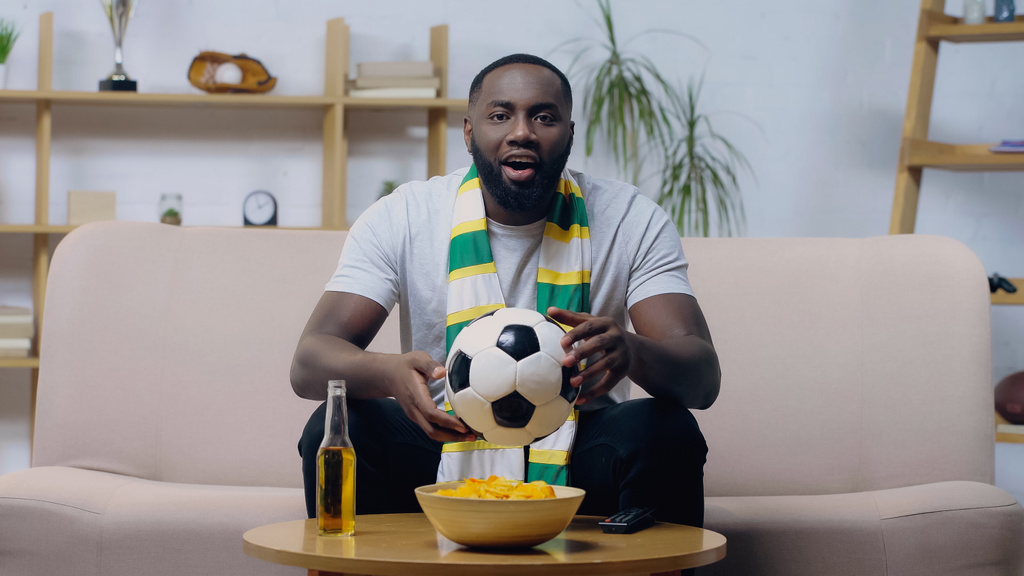 emocionado hombre afroamericano sosteniendo pelota de fútbol mientras mira campeonato de fútbol cerca de cerveza y patatas fritas - Foto, Imagen
