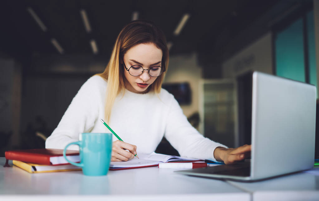 Young female concentrated student in white jumper and eyeglasses sitting at table  with laptop and taking notes in notebook in cafe on blurred background - Photo, Image