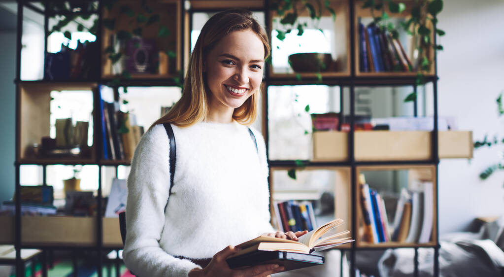 Usmívající se blond dospívající stojící ve dne v knihovně světla a hledající zajímavou literaturu v moderním knihkupectví při nošení bílého svetru a batohu při pohledu do kamery - Fotografie, Obrázek