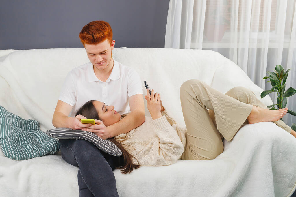 Dos personas cariñosas relajantes fin de semana en casa en el sofá sosteniendo en las manos los teléfonos inteligentes y charlando. - Foto, imagen