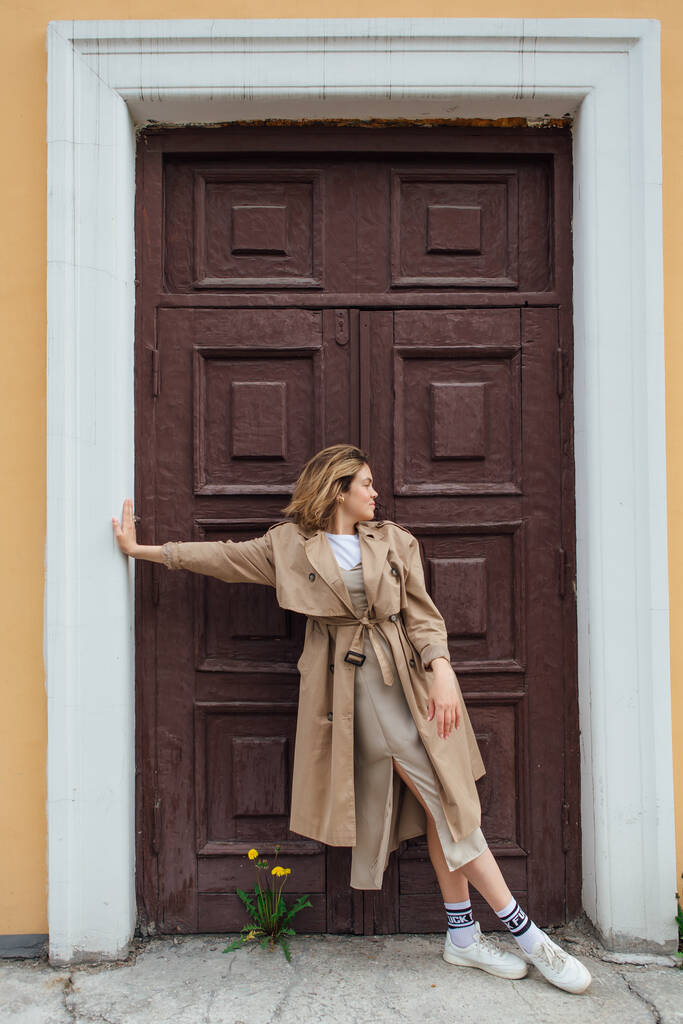 Mladá usmívající se tisíciletá žena s divokými vlasy oblečená v podzimním kabátě pózující u dveří staré budovy. - Fotografie, Obrázek