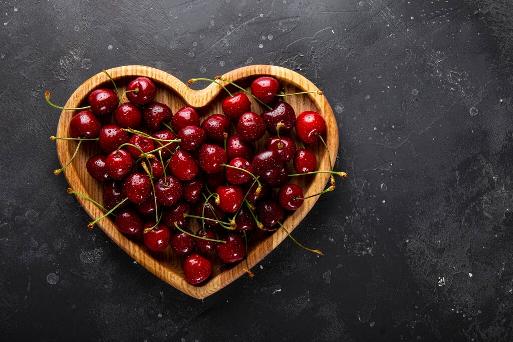 Cerezas maduras y fresas en un plato de madera en forma de corazón y fondo gris oscuro, tarjeta de San Valentín con fruta - Foto, imagen