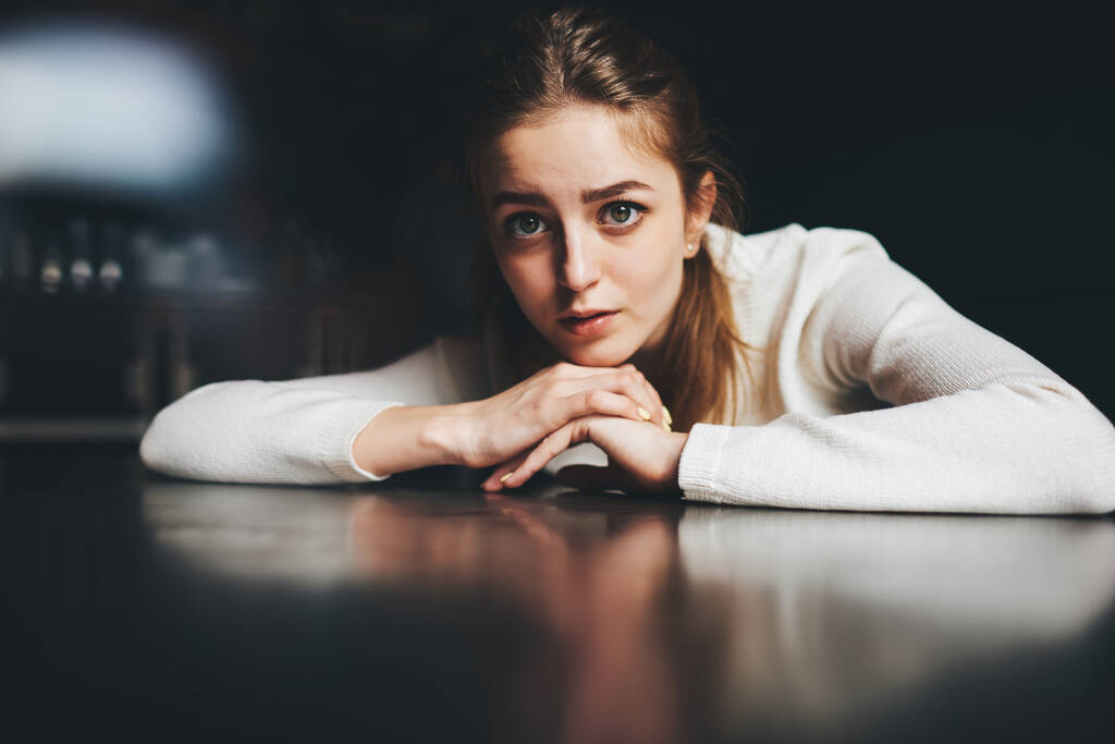 Jong pensive en verdrietig vrouw in witte trui zitten aan tafel zetten hoofd op handen en kijken naar de camera in de kamer - Foto, afbeelding