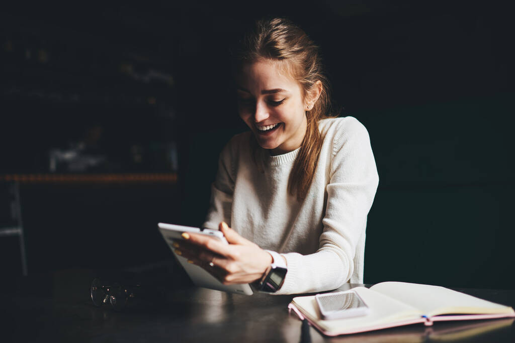 Szczęśliwa młoda kobieta w białych ubraniach z zębate uśmiech siedzi przy stole z otwartym notatnikiem podczas rozmowy wideo na tablecie w nocy - Zdjęcie, obraz