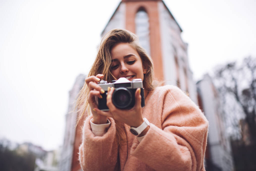 Bajo ángulo de la joven fotógrafa en ropa de abrigo con sonrisa dentada sosteniendo la cámara antigua mientras toma fotos en la calle - Foto, imagen