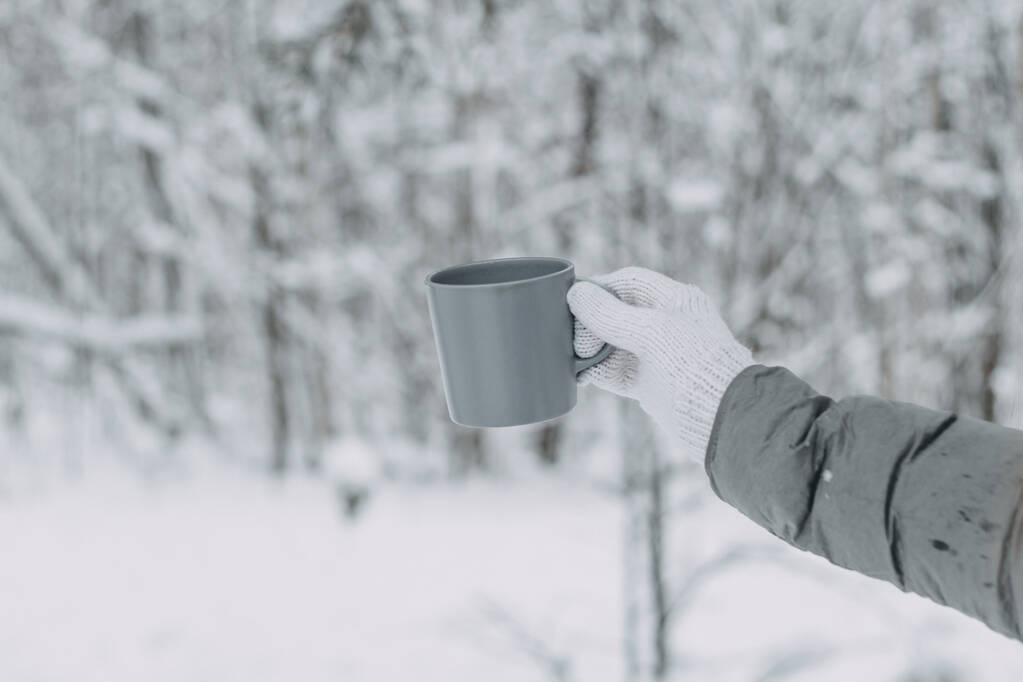Una mano en un guante de lana blanco y una chaqueta gris sostiene una taza gris sobre el fondo de un hermoso bosque cubierto de nieve. - Foto, imagen