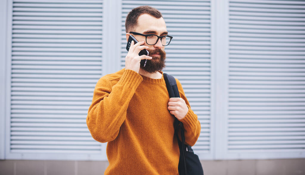 自信のある髭の男の広い角度離れて携帯電話の呼び出しを作るためにスマートフォンを使用して街の通りを歩いて自由な時間を過ごす - 写真・画像