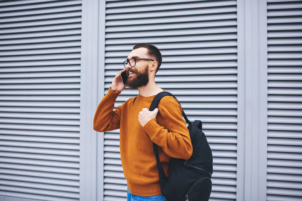 Sivukuva iloinen mies reppu ja silmälasit katselee pois ottaa keskustelun matkapuhelimeen viettäessään aikaa kadulla - Valokuva, kuva