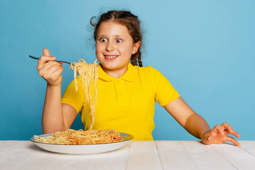 Roztomilé mladé dívky, emotivní dítě jíst lahodné italské těstoviny izolované na modrém pozadí studia. Světový den těstovin - Fotografie, Obrázek