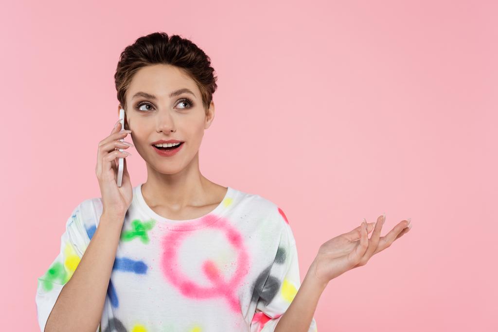 mujer feliz con el pelo corto apuntando con la mano mientras habla en el teléfono móvil aislado en rosa - Foto, imagen