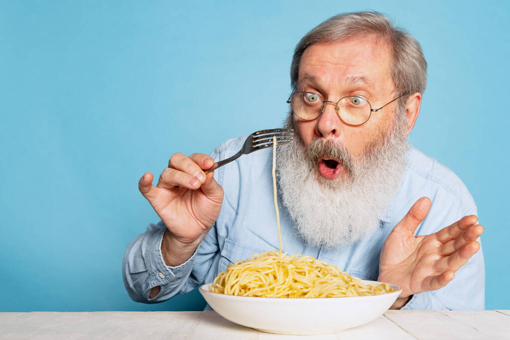 Šokovaný šedovousý muž je překvapen velkou porcí nudlí, těstovin, izolovaných na modrém pozadí studia. - Fotografie, Obrázek