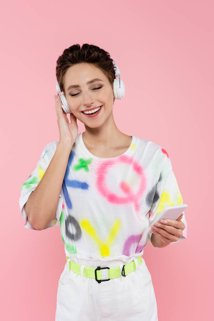 heureux jeune femme avec téléphone portable réglage casque tout en écoutant de la musique isolée sur rose - Photo, image