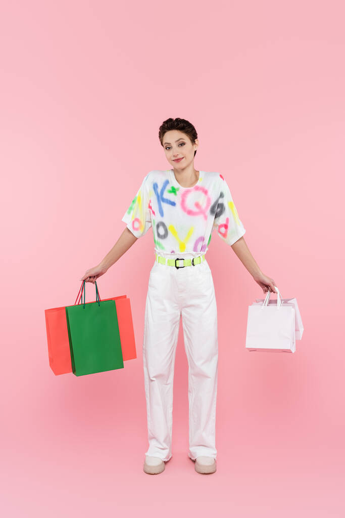 visão de comprimento total da mulher sorridente com sacos de compras multicoloridos olhando para a câmera em rosa - Foto, Imagem