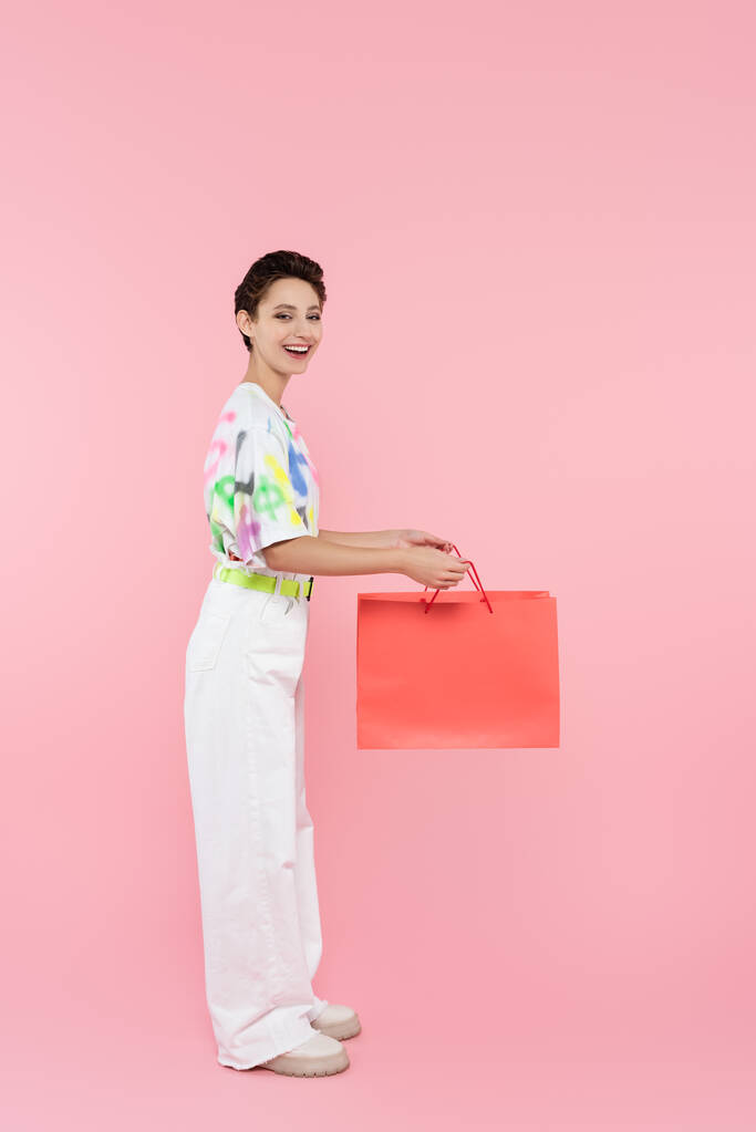 vista completa de mujer feliz en pantalones blancos sosteniendo bolsa de compras en rosa - Foto, Imagen