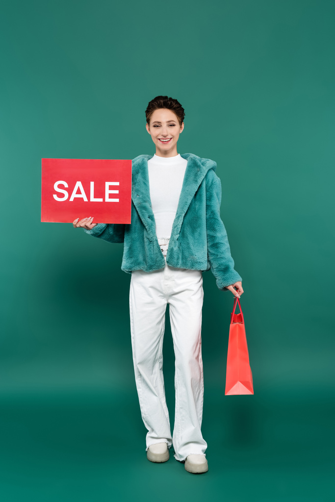 mujer feliz en chaqueta de piel sintética y pantalones blancos con bolsa de compras y tarjeta roja con letras de venta en verde - Foto, Imagen