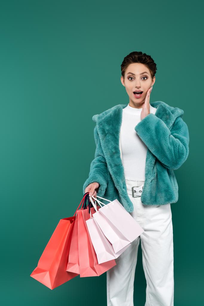 mulher espantada em roupas da moda tocando rosto enquanto segurando sacos de compras multicoloridos isolados no verde - Foto, Imagem