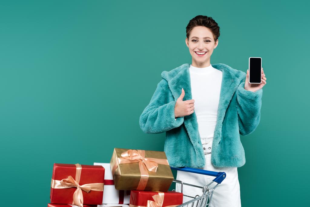 mujer sonriente mostrando el pulgar hacia arriba mientras sostiene el teléfono inteligente con pantalla en blanco cerca del carrito de compras con regalos aislados en verde - Foto, imagen