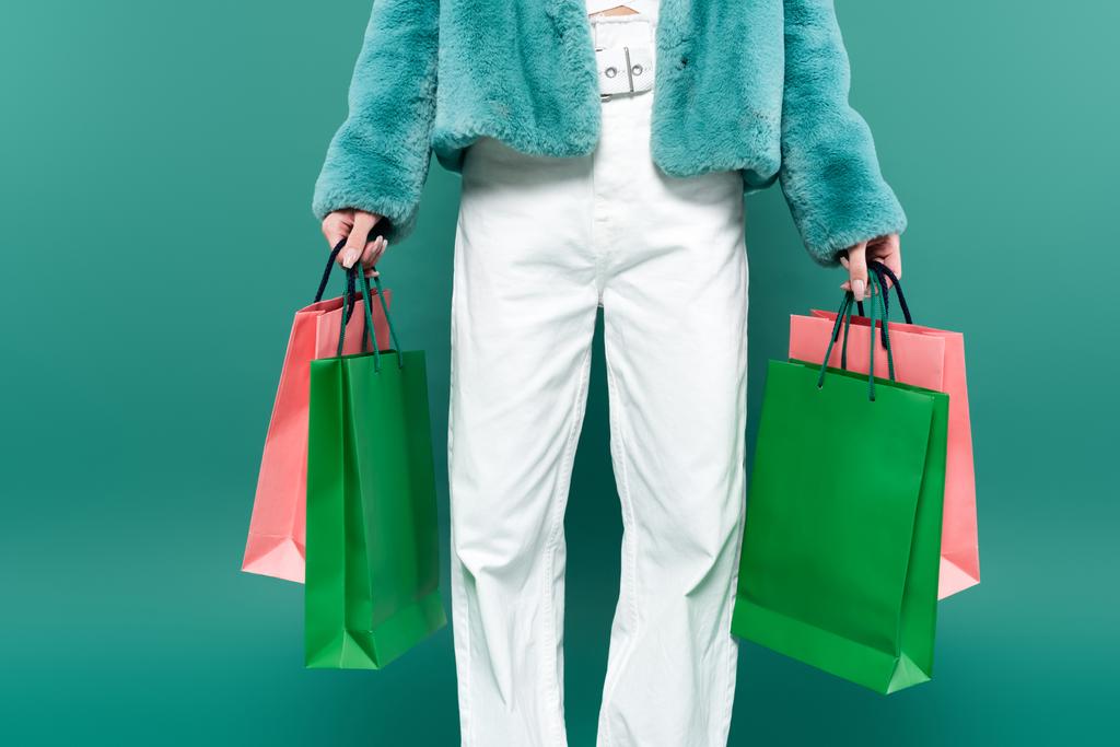 częściowy widok kobiety w sztucznej kurtce futerkowej i białych spodniach posiadających wielobarwne torby na zakupy izolowane na zielono - Zdjęcie, obraz