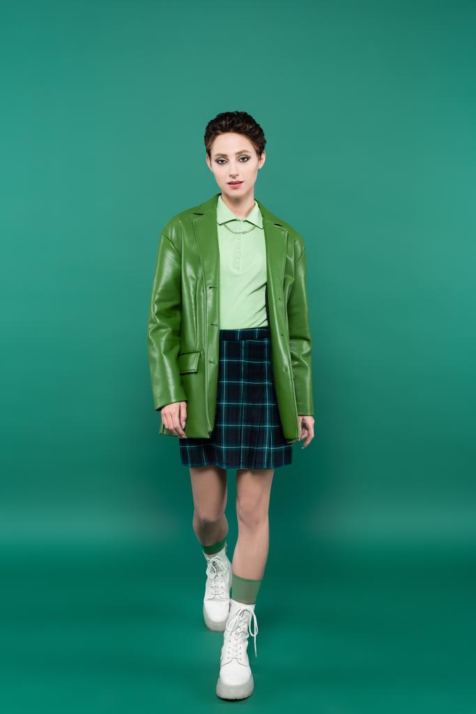 vista completa de la mujer de moda en chaqueta de cuero, falda a cuadros y botas blancas atadas caminando sobre verde - Foto, imagen