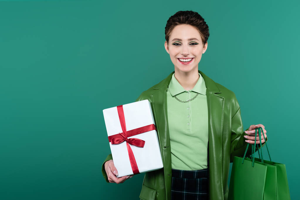 jeune et élégante femme tenant boîte cadeau et sacs à provisions tout en souriant à la caméra isolée sur vert - Photo, image