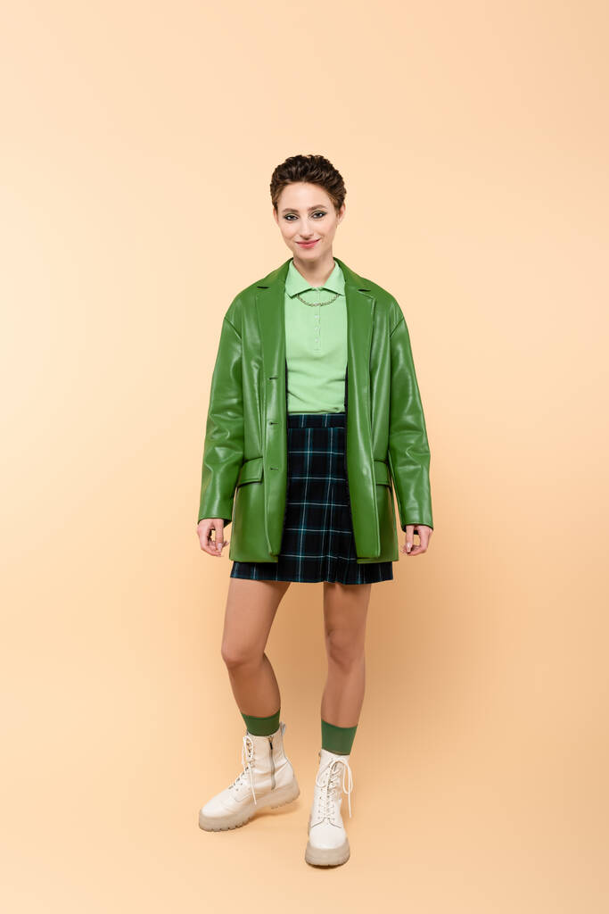 full length view of positive woman in green jacket, plaid skirt and white lace-up boots on beige - Foto, Imagem