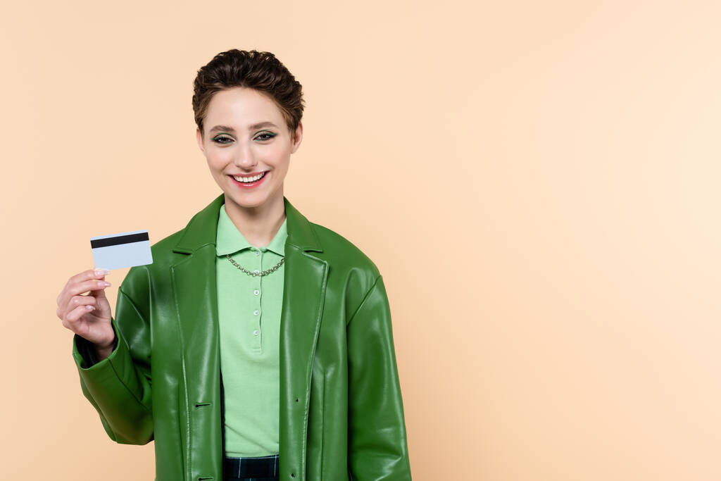 freudige brünette Frau mit Kreditkarte blickt isoliert in die Kamera auf beige - Foto, Bild