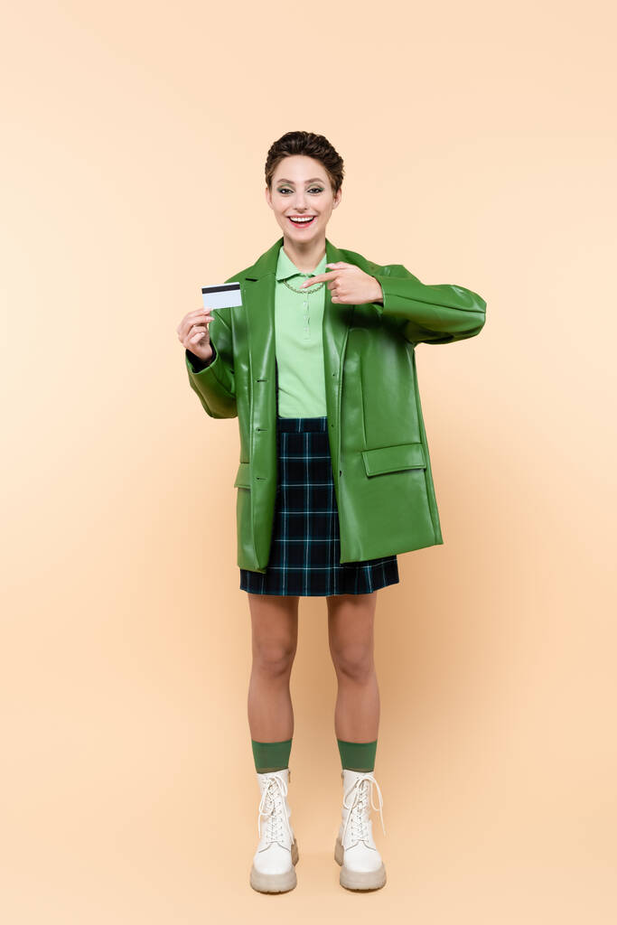 πλήρους μήκους άποψη της χαρούμενης γυναίκας σε καρό φούστα και πράσινο σακάκι που δείχνει προς την πιστωτική κάρτα σε μπεζ - Φωτογραφία, εικόνα
