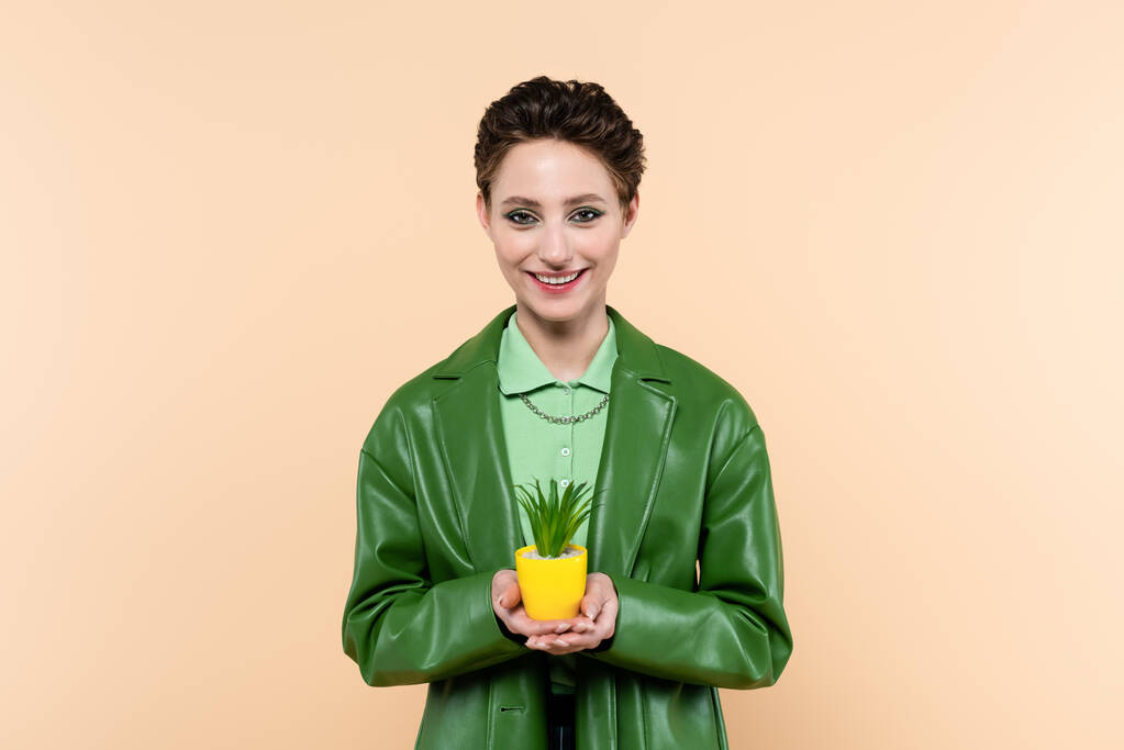 ευτυχισμένη γυναίκα σε πράσινο σακάκι εκμετάλλευση φυτό σε κίτρινο γλάστρα απομονωμένη σε μπεζ - Φωτογραφία, εικόνα