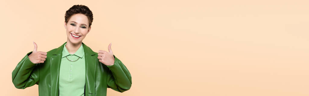 Yeşil renkli ceketli neşeli genç kadın, başparmaklarını bej ve pankartta gösteriyor. - Fotoğraf, Görsel