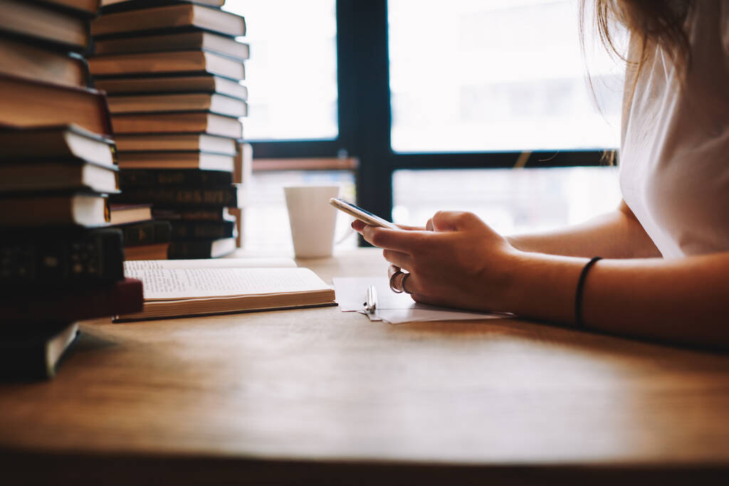 Vista lateral de una bibliotecaria irreconocible navegando por un teléfono inteligente sentado en una mesa de madera con muchos libros y una taza con bebida caliente - Foto, Imagen