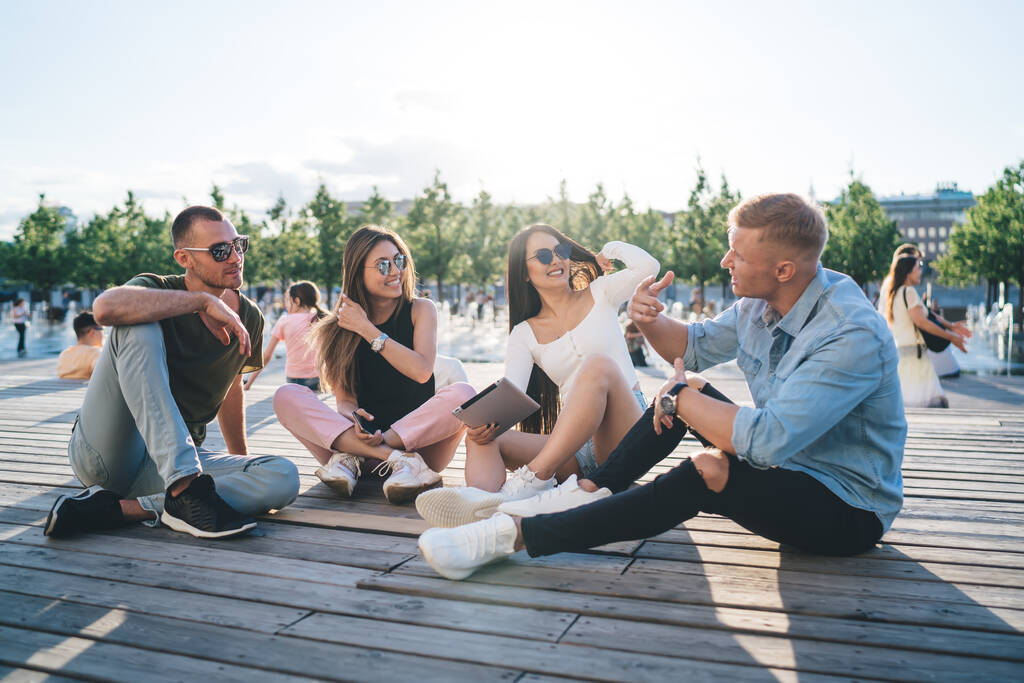 Grupa młodych szczęśliwych przyjaciół rozmawia ze sobą siedząc na pięknym nabrzeżu w pogodny letni dzień i ciesząc się tym czasem - Zdjęcie, obraz