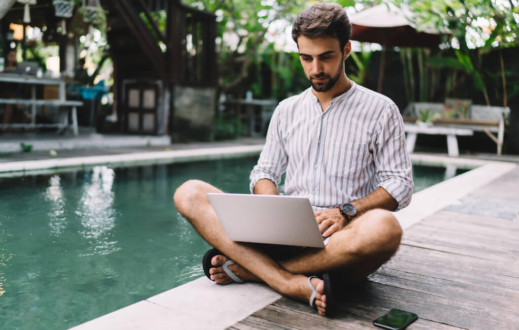 Młody przedsiębiorca w letniej koszuli i japonkach siedzących na basenie i korzystających z laptopa do prowadzenia internetowego projektu biznesowego w cichym podwórku ośrodka - Zdjęcie, obraz