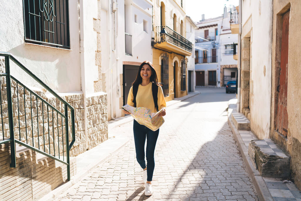 Full body of cheerful ethnic female in casual clothes and backpack holding map and walking on pavement while looking at camera - Photo, Image