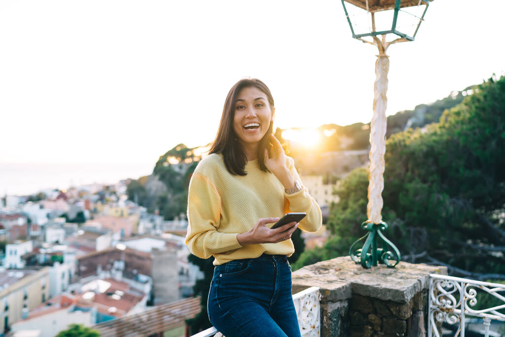 Happy Asian brunette female in casual clothes with toothy smile sitting on stone parapet and browsing smartphone while looking at camera - Photo, Image