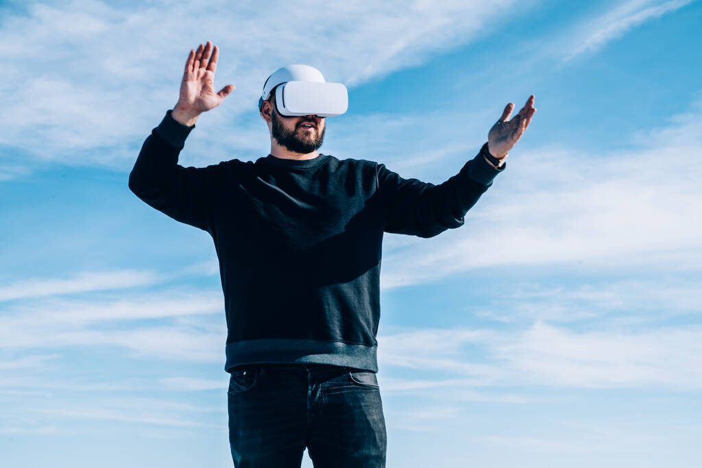 Von unten staunende männliche Spieler in Freizeitkleidung mit Virtual-Reality-Brille, während sie bei trübem Tag mit erhobenen Armen draußen stehen - Foto, Bild