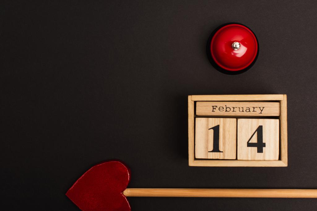 вид стрелы в форме сердца рядом с деревянными кубиками с 14 февраля надписью и колоколом на черном  - Фото, изображение