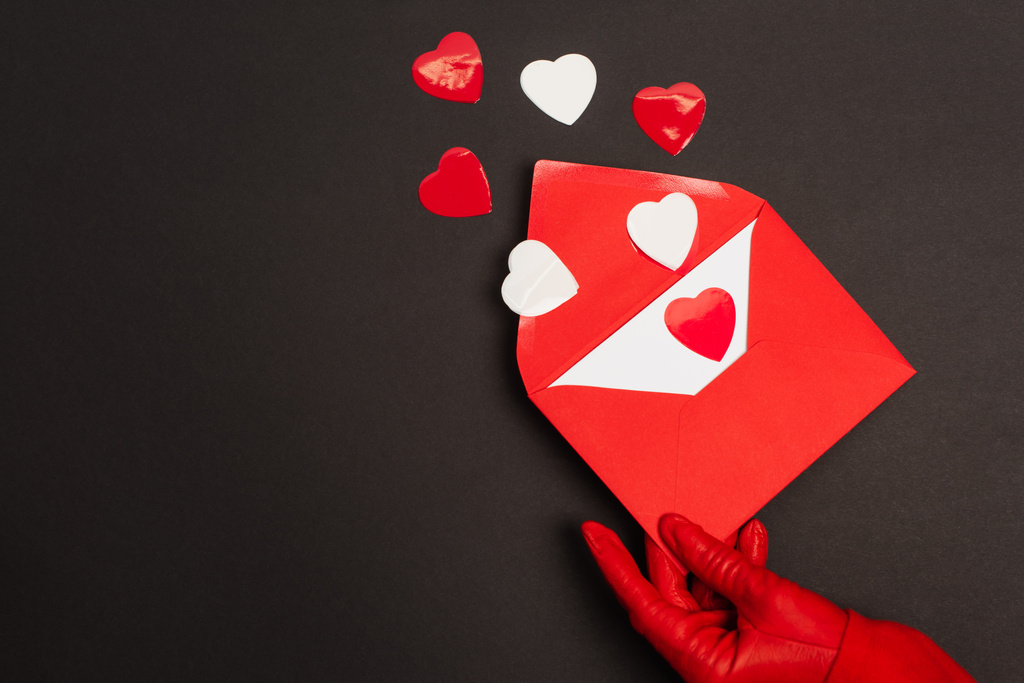 vue recadrée de la personne en gant rouge tenant enveloppe avec lettre d'amour et des coeurs coupés en papier sur noir  - Photo, image
