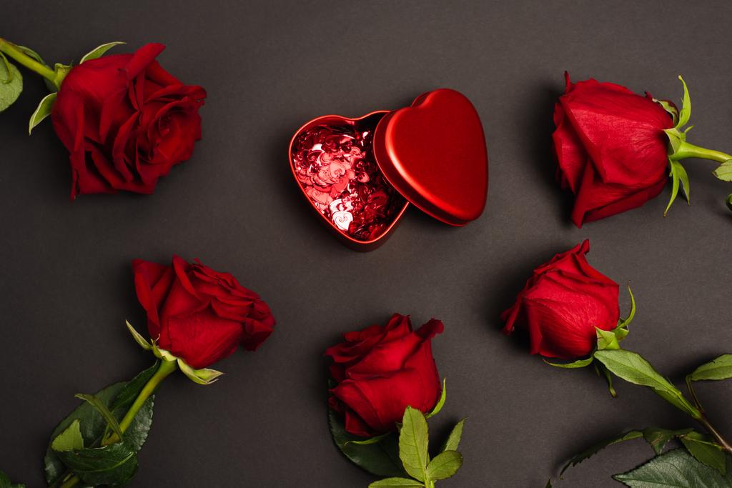 πάνω όψη του μεταλλικού κουτιού με κόκκινες καρδιές κομφετί κοντά σε τριαντάφυλλα σε μαύρο - Φωτογραφία, εικόνα
