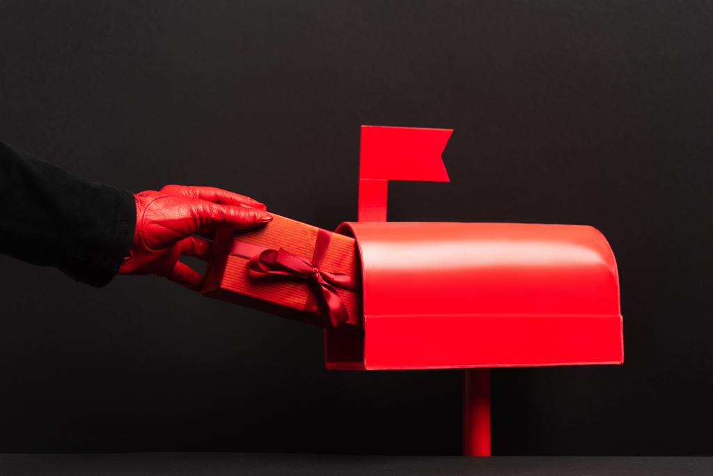 μερική άποψη του προσώπου με κόκκινο γάντι βάζοντας παρόν σε ταχυδρομική θυρίδα σε μαύρο  - Φωτογραφία, εικόνα